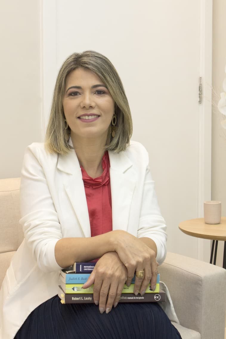 Valquiria Fernandes, psicóloga clínica, sorrindo sentada no sofá de seu consultório em Perdizes, São Paulo, de blazer branco, segurando livros de psicologia